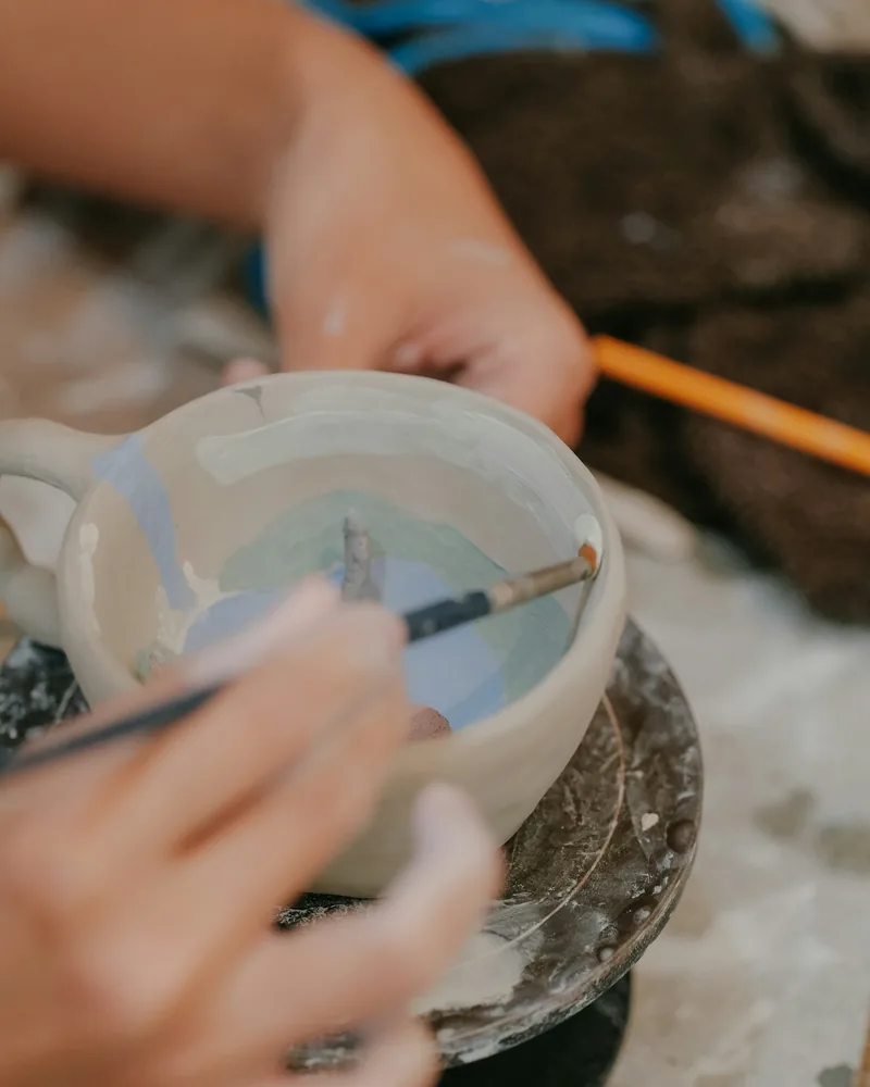 Pincel aplicando esmalte azul y verde sobre el interior de una taza bizcochada en una mesa de taller